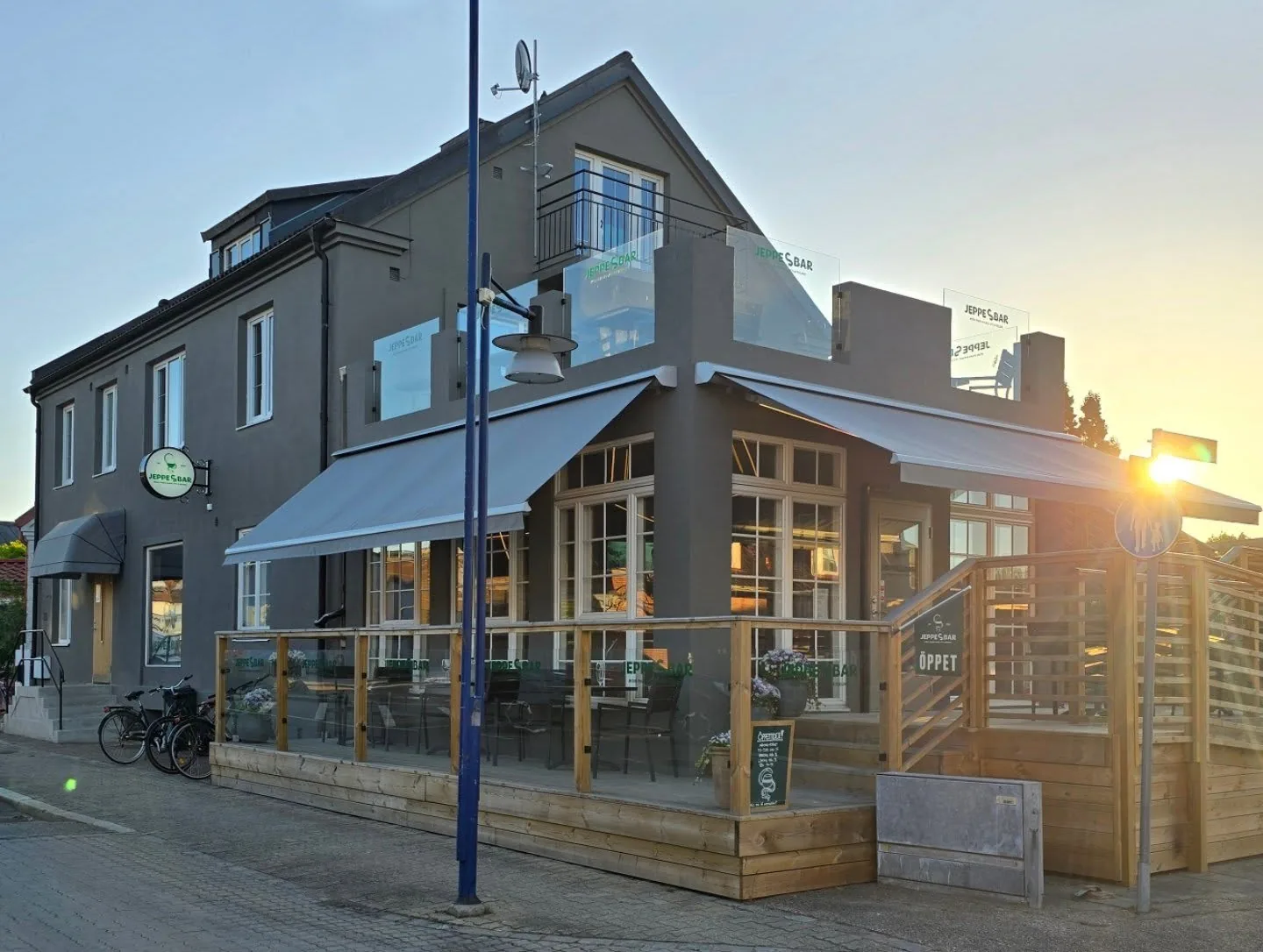Hörnhuset i mörkgrå puts med den inglasade verandan, grå markiser och trädäcket runt hörnet i kvällssol