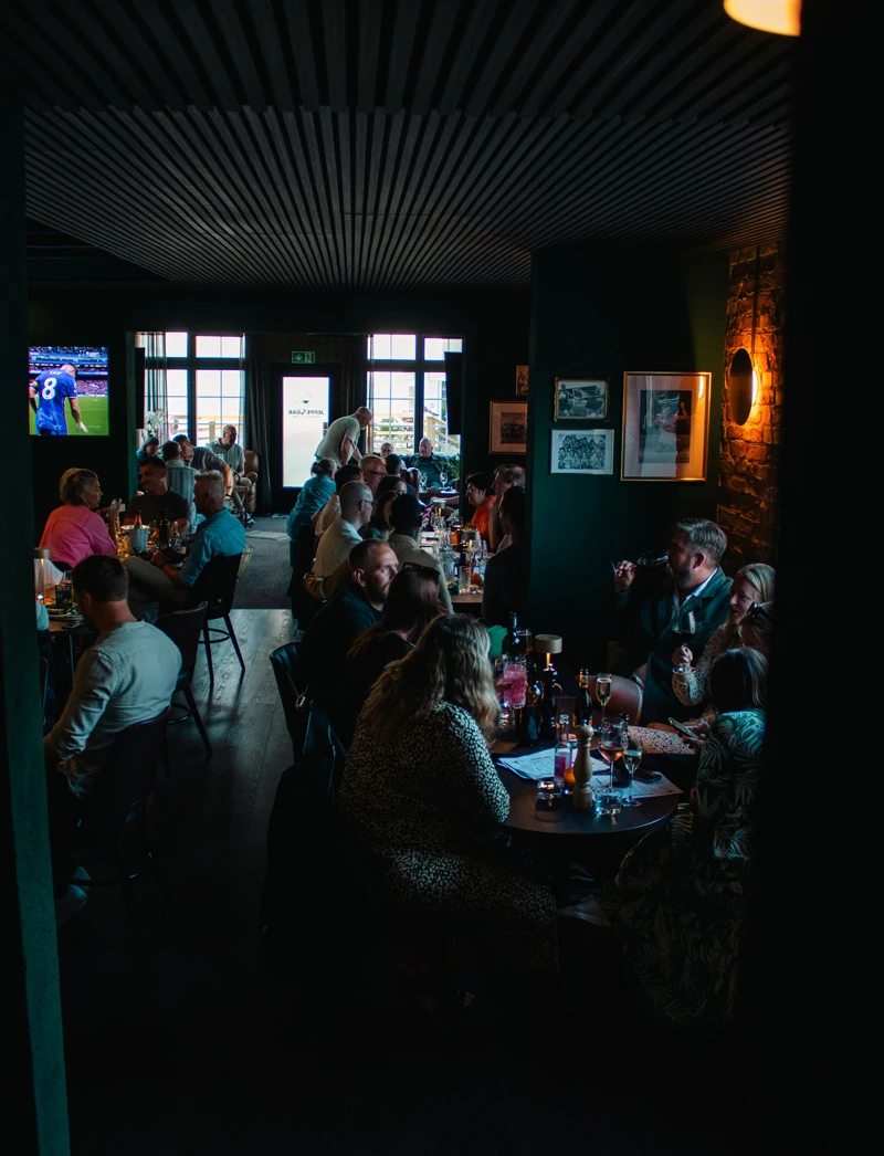 Fullsatt matsal en kväll: gäster vid långa bord under gröna väggar, fotboll på tv:n och fönsterraden mot gatan i fonden